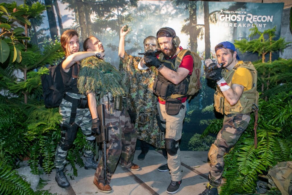 Cinq personnes déguisées en aventurier : quatre hommes et une femme qui prennent la pose devant un photocall Ghost Recon qui ressemble à de la jungle.