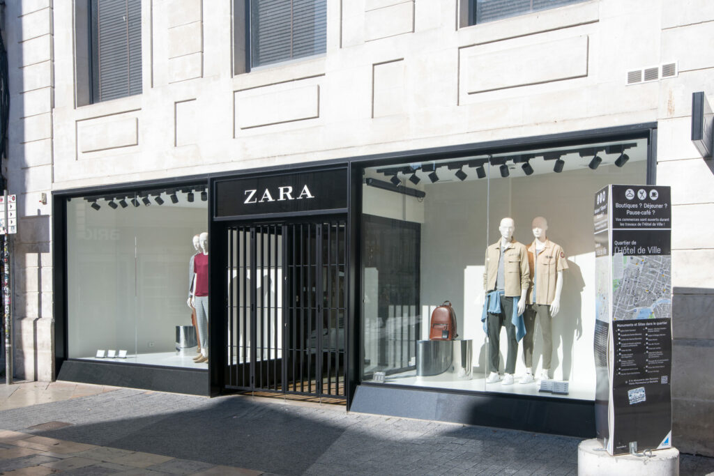 Une devanture noire de boutique Zara, avec trois mannequins dans la vitrine habillés avec des vêtements d'été.