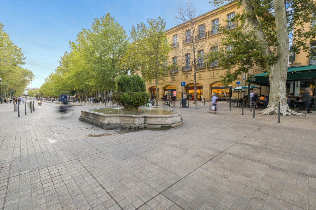 Place en pleine ville avec en son centre une fontaine et en arrière plan une rangée d'arbres placée devant un immeuble jaune.