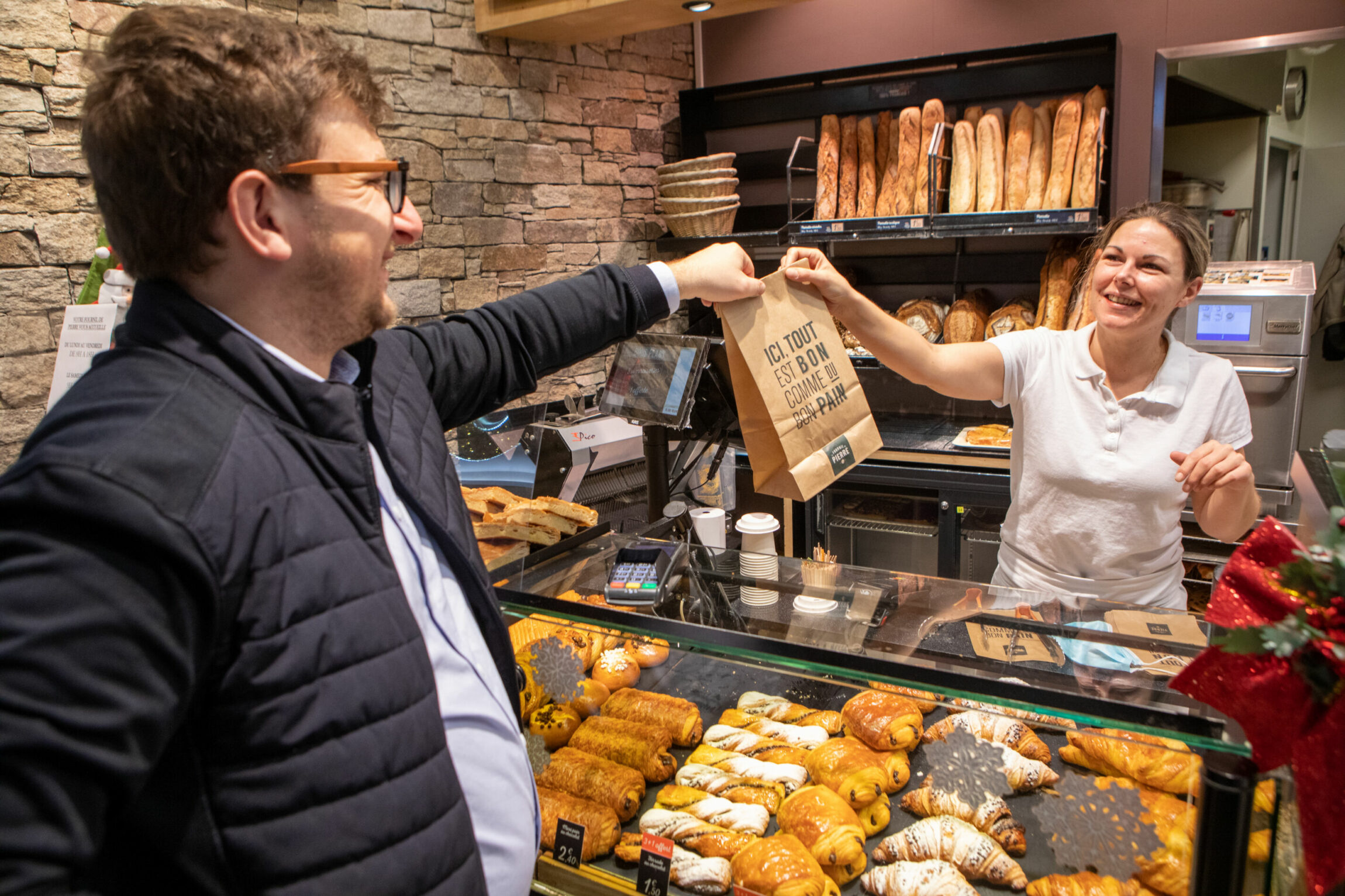 Un client dans une boulangerie prenant son sac de viennoiserie que le vendeuse lui tend.